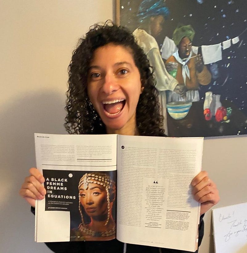 Chanda smiling while holding open a magazine featuring text that reads “A Black Femme Dreams in Equations” in front of an art piece featuring black women.