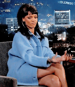 Rihanna in a blue jacket on a talk show making the schmoney sign with her fingers and nails.