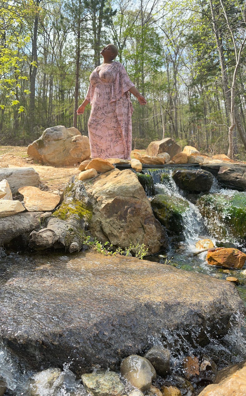 Keila stands on rocks in the center of a stream, face turned upward toward the sun.