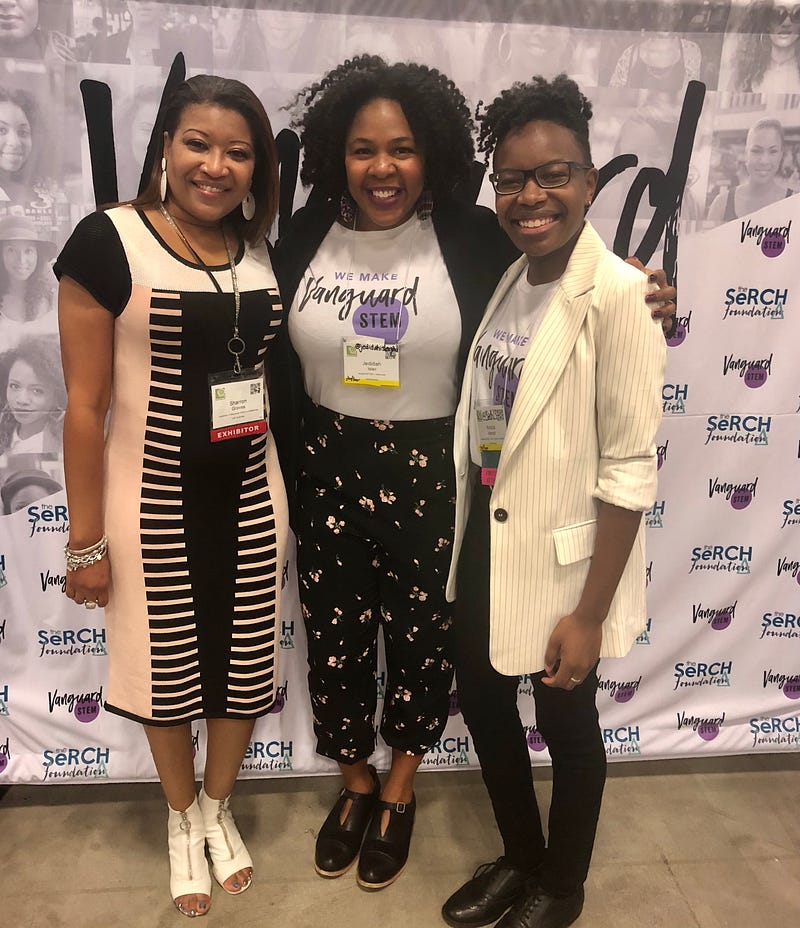Sharron (left) stands in front of the #VanguardSTEM back drop. To her right are Jedidah Isler, Founder and CEO of #VS and Anicca Harriot, #VS Chief of Community Development. All are smiling toward the camera.