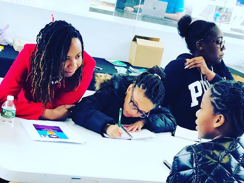 Malika leaning over a table in a red top chatting with 3 young Black girls, all in black, as one is taking notes.