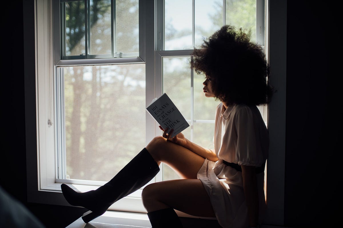 A femme-presenting person reading a book on the ledge of a window.