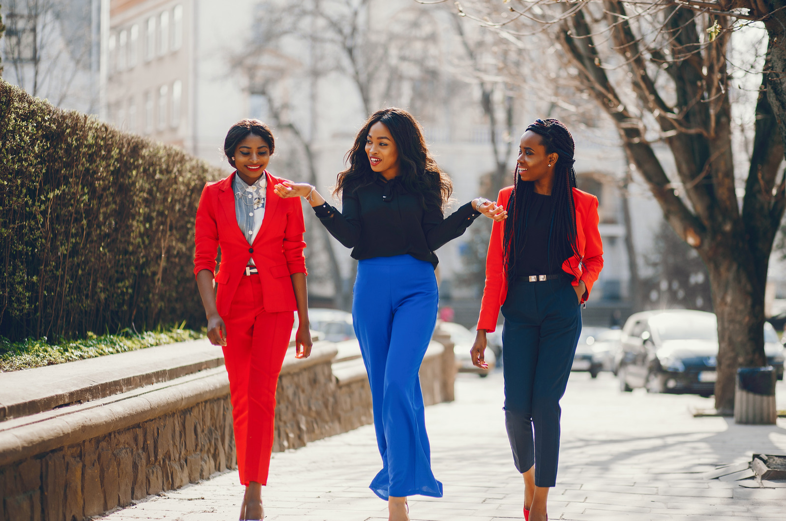Three women of color are walking on a sidewalk. On the far left is a woman in a red suit with her hair pulled back, center is a woman in a black blouse and cobalt blue pants gesturing with her hands, on the right is a woman dressed in all black with a red blazer; her hair is braided and pulled back.