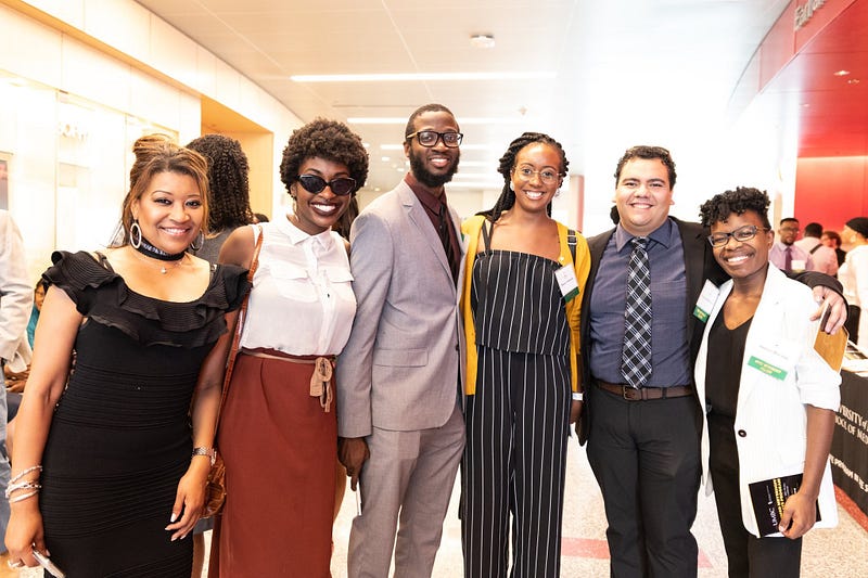 Sharron (left) smiles at the camera. Standing to her right also smiling are five doctoral students of color of various gender identities, all dressed in business attire.