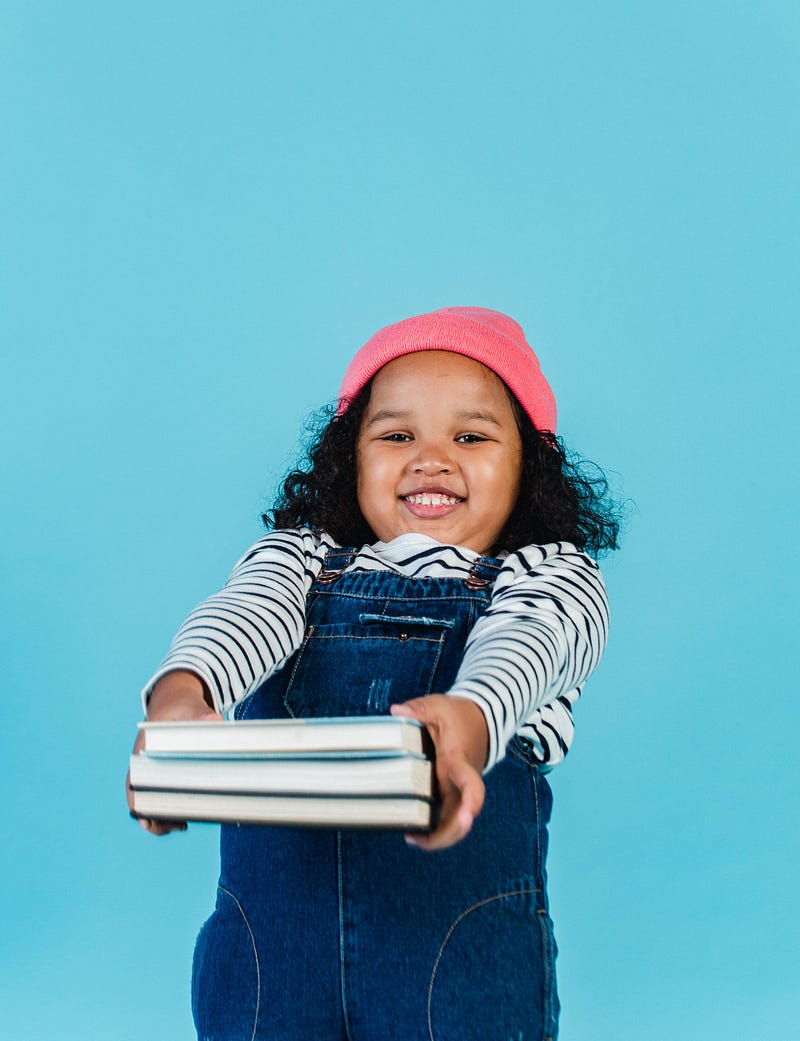 A young girl holding out two books with a pink hat, blue jean overalls and a striped shirt.