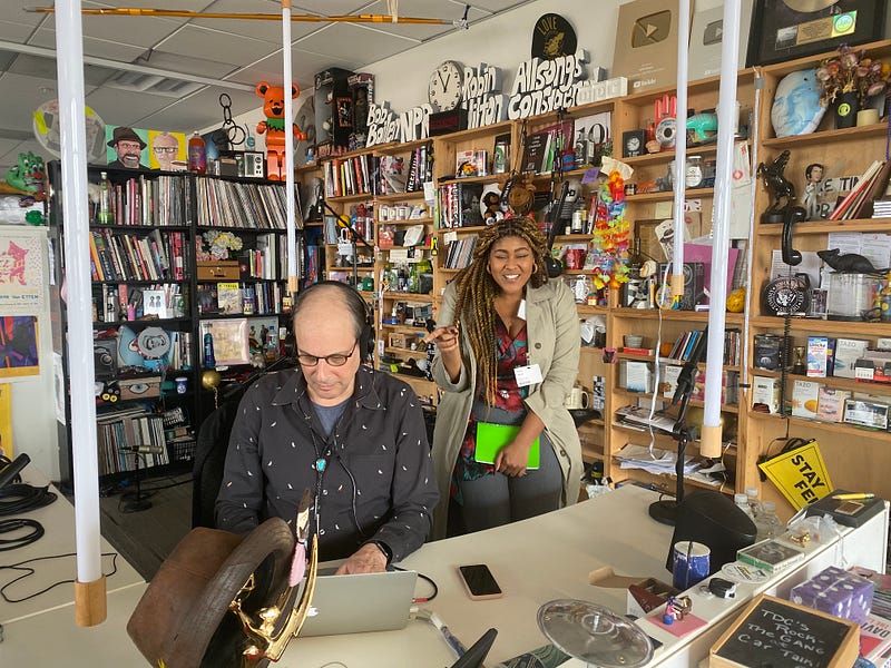 Keila sits at the NPR Tiny Desk set, just behind Bob Boilen, pointing at him and smiling as he works.
