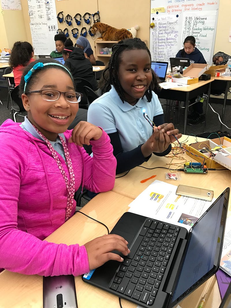 Two girls sit side by side smiling at the camera. One is coding on a laptop while the other tinkers with a circuit board.